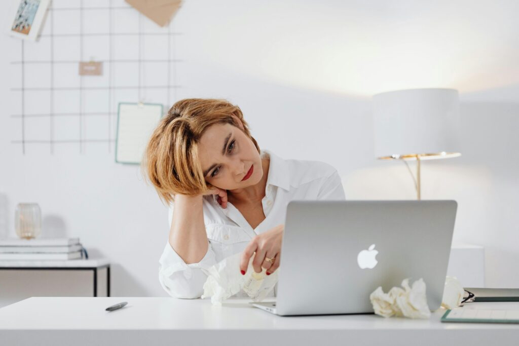 A tired woman in a white shirt sitting at a desk while using a laptop, appearing weary and bored.