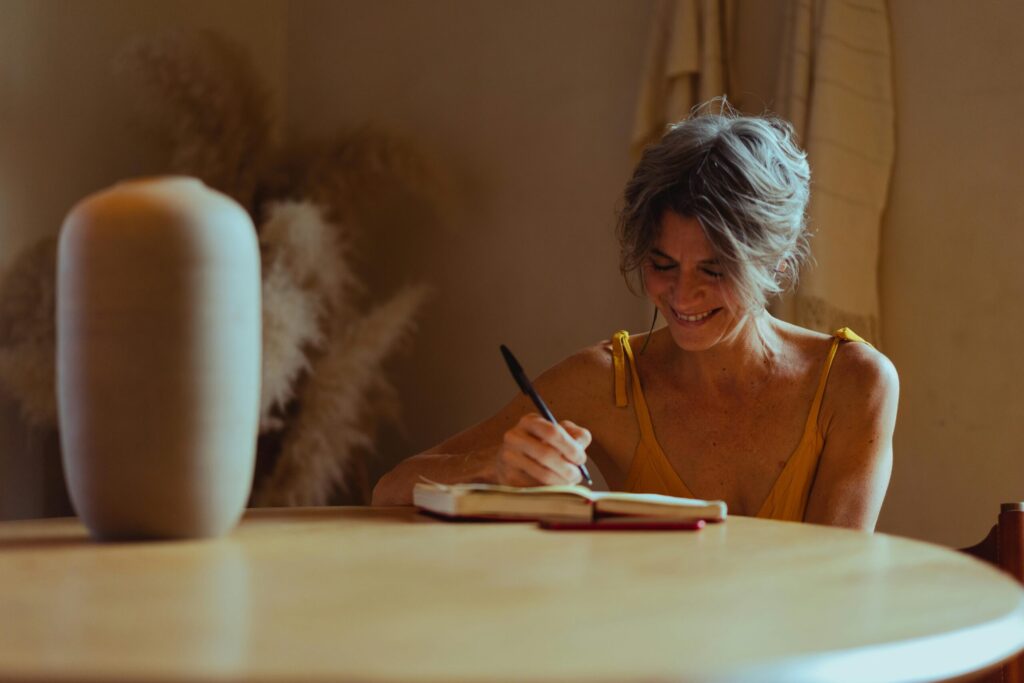 A senior woman sitting at a table, journaling happily in her cozy home office.